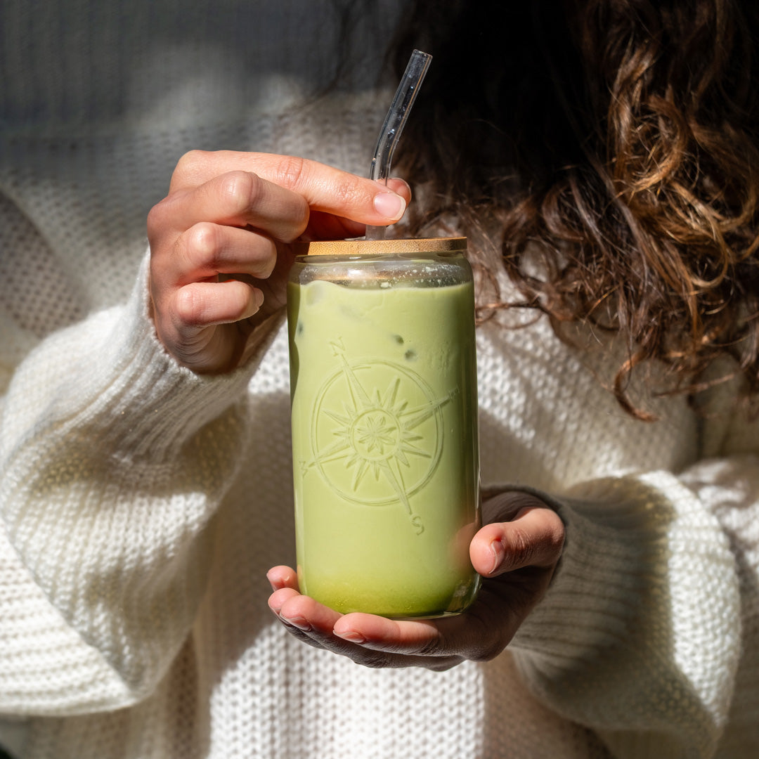 A person in a white knit sweater holds a Matcha Latte (Kit) in a clear glass jar with a compass design, wooden lid, and glass straw. Sunlight highlights their hands and the vibrant ceremonial matcha swirling inside.